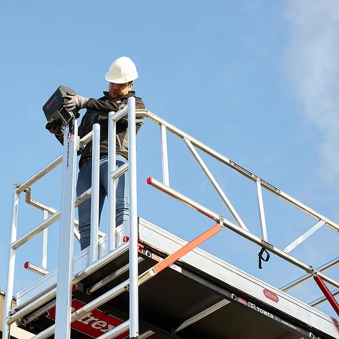 Altrex Rollgerüst Shuttle Basisset mit Winde 3,20m Plattformhöhe 5,20m Arbeitshöhe (kein Ge (Arbeitshöhe: 5.200 m, Belastbarkeit Plattform: 30 kg) -  - Altrex RS Tower 5 Fahrgerüst mit Arbeiter auf der Plattform. Silberner Aluminiumrahmen, schwarze Fiber-Decks und rote Akzente.