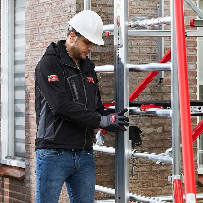Altrex Rollgerüst Shuttle Basisset mit Winde 3,20m Plattformhöhe 5,20m Arbeitshöhe (kein Ge (Arbeitshöhe: 5.200 m, Belastbarkeit Plattform: 30 kg) -  - Mann in Schutzausrüstung montiert ein Altrex Gerüst. Das Gerüst hat silberne Metallrahmen und rote Akzente.