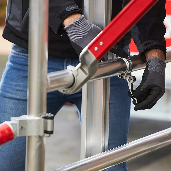 Altrex Rollgerüst Shuttle Basisset mit Winde 3,20m Plattformhöhe 5,20m Arbeitshöhe (kein Ge (Arbeitshöhe: 5.200 m, Belastbarkeit Plattform: 30 kg) -  - Behandschuhte Hände verbinden silberne Gerüstrohre mit einer Schnellspannklemme und einer roten Stange.