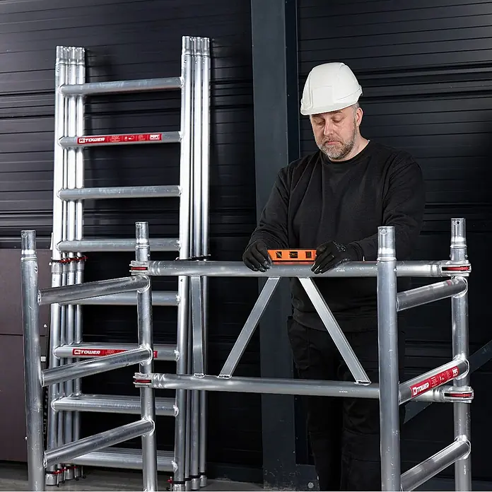 Altrex Rollgerüst MiTower 1-Personen-Gerüst Aluminium mit Holz-Plattform 0,75x1,27m 5,20m AH - Arbeitshöhe: 5.200 m - Silberne Aluminium-Gerüstteile, von einem Arbeiter mit Schutzhelm und Handschuhen, der eine Wasserwaage benutzt, auf Richtigkeit geprüft.