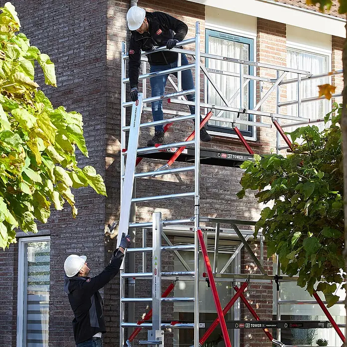 Altrex Rollgerüst Shuttle Basisset mit Winde 3,20m Plattformhöhe 5,20m Arbeitshöhe (kein Ge (Arbeitshöhe: 5.200 m, Belastbarkeit Plattform: 30 kg) -  - Zwei Arbeiter montieren ein silbernes Altrex RS Tower 5 Gerüst mit roten Streben an einem Backsteinhaus.
