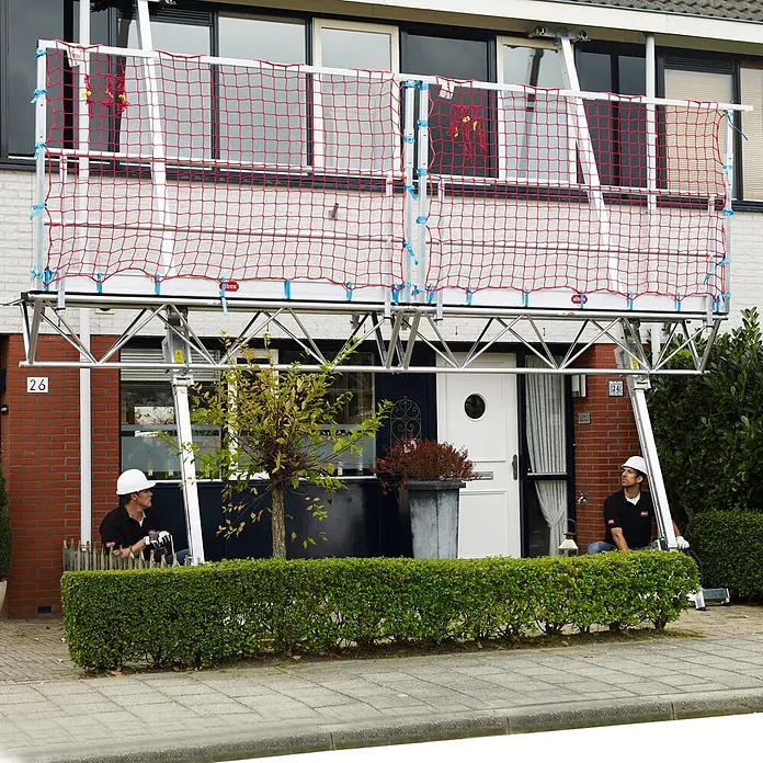 Altrex Rollgerüst Modulare Dreiecksbühne Absturzsicherung mit Fangnetzen für geneigte Däch -  - Modulares Aluminiumgerüst mit rotem Schutznetz vor einem Haus für Höhenarbeiten. Zwei Arbeiter beobachten.