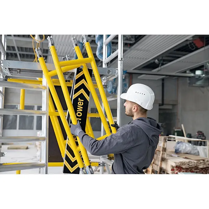 Munk Rollgerüst FlexxTower Ein-Personen-Gerüst 4,10 AH - Arbeitshöhe: 4.100 m - Arbeiter montiert gelbe Gerüstturmteile mit schwarzer gemusterter Platte.
