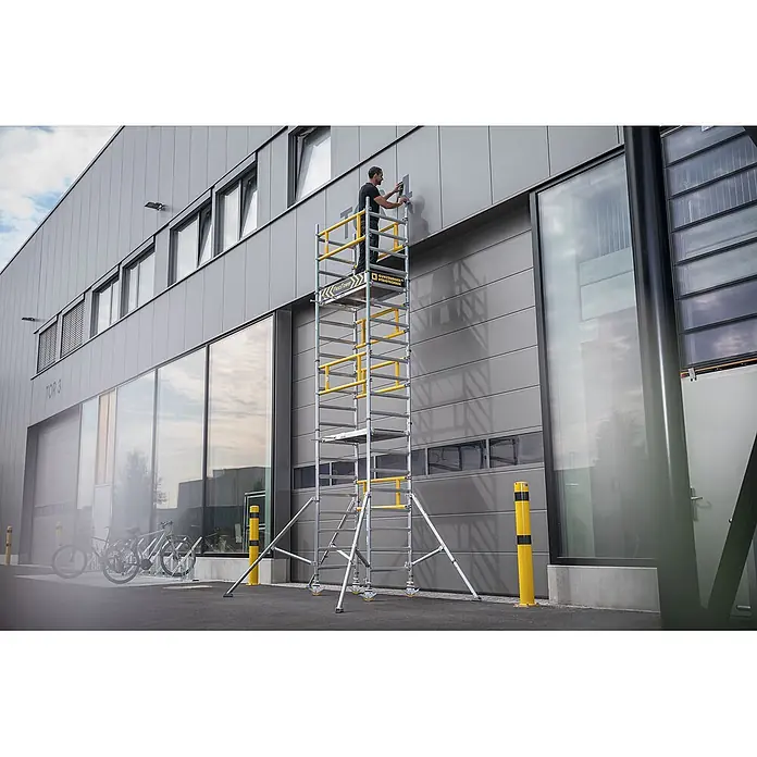 Munk Rollgerüst FlexxTower Ein-Personen-Gerüst 4,10 AH - Arbeitshöhe: 4.100 m - Ein Mann auf einem hohen, mobilen Aluminium-Rollgerüst mit gelben Sicherheitsgeländern arbeitet an einer Gebäudewand.