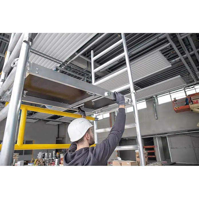 Munk Rollgerüst FlexxTower Ein-Personen-Gerüst 4,10 AH - Arbeitshöhe: 4.100 m - Arbeiter montiert eine moduläre Aluminium-Gerüstplattform mit dunkler Holzoberfläche.