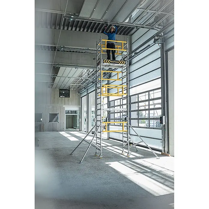 Munk Rollgerüst FlexxTower Ein-Personen-Gerüst 4,10 AH - Arbeitshöhe: 4.100 m - FlexxTower Rollgerüst, silber mit gelben Akzenten, wird von einem Arbeiter in einem großen Gebäude verwendet.