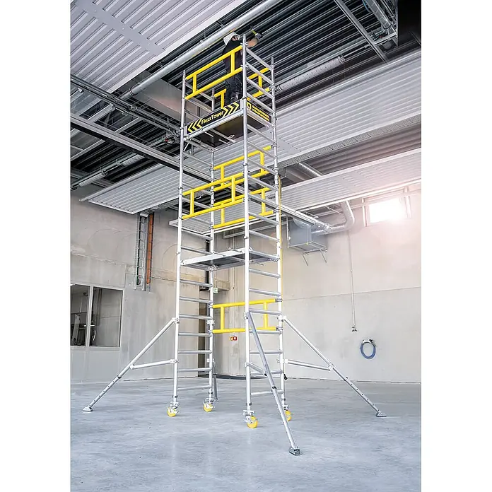 Munk Rollgerüst FlexxTower Ein-Personen-Gerüst 4,10 AH - Arbeitshöhe: 4.100 m - Mobiles Aluminium-Rollgerüst, silber mit gelben Geländern, wird von einem Arbeiter in einem Gebäude verwendet.