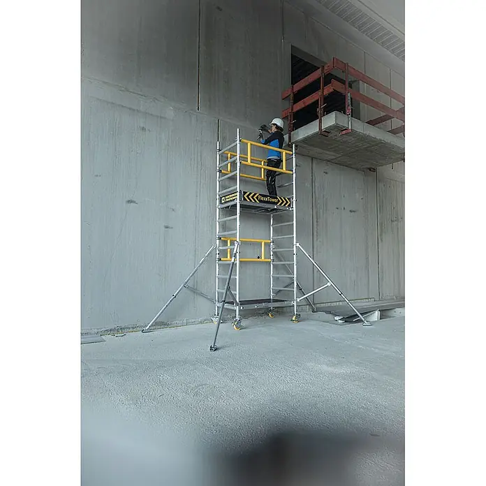 Munk Rollgerüst FlexxTower Ein-Personen-Gerüst 4,10 AH - Arbeitshöhe: 4.100 m - Silberner Rollgerüstturm mit gelben Akzenten, von einem Arbeiter an einer Betonwand genutzt.