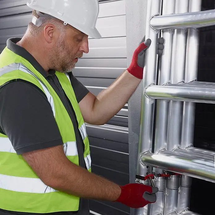 Altrex Rollgerüst MiTower PLUS Fahrgerüst 1-Person-Aufbau Aluminium mit breiter Fiber-Deck P - Arbeitshöhe: 5.200 m - Mann in Schutzkleidung justiert ein silbernes Metallrohrsystem.