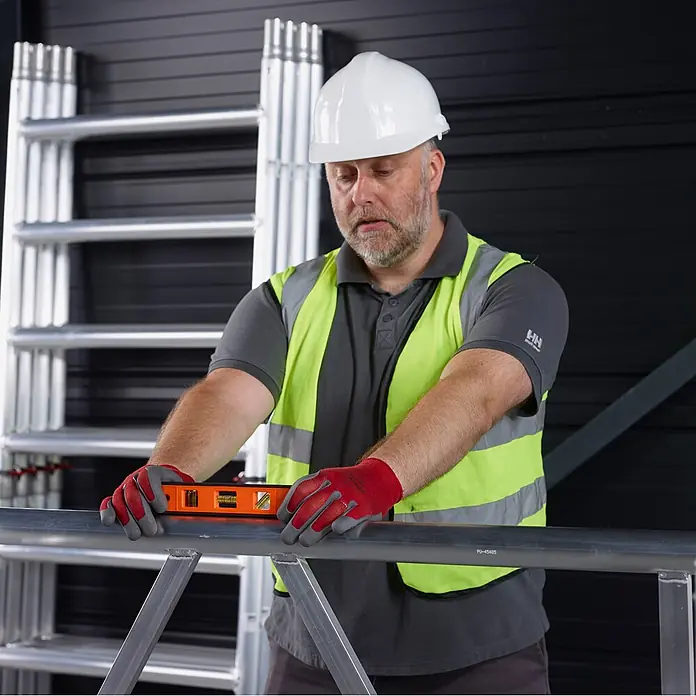 Altrex Rollgerüst MiTower PLUS Fahrgerüst 1-Person-Aufbau Aluminium mit breiter Fiber-Deck P - Arbeitshöhe: 5.200 m - Ein Bauarbeiter in Helm und Warnweste prüft ein silbernes Gerüstrohr mit einer orange-schwarzen Wasserwaage.