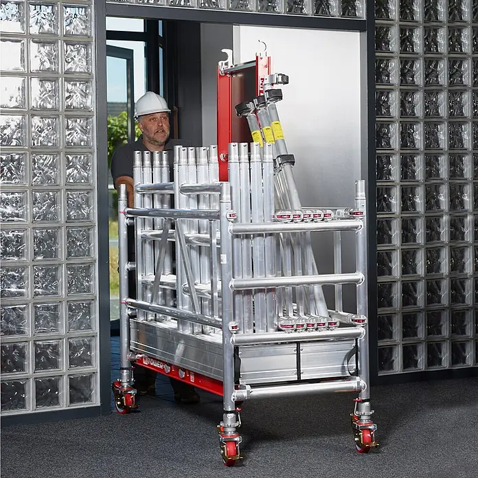 Altrex Rollgerüst MiTower PLUS Fahrgerüst 1-Person-Aufbau Aluminium mit breiter Fiber-Deck P - Arbeitshöhe: 5.200 m - Ein Mann mit Helm schiebt ein mobiles Aluminium-Gerüst mit roten Rollen durch eine Tür, was seine Tragbarkeit hervorhebt.