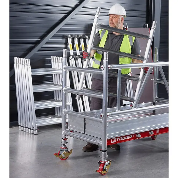 Altrex Rollgerüst MiTower PLUS Fahrgerüst 1-Person-Aufbau Aluminium mit breiter Fiber-Deck P - Arbeitshöhe: 5.200 m - Ein Arbeiter mit Schutzhelm und Warnweste montiert ein silbernes Aluminium-Rollgerüst mit rotem Unterbau und Lenkrollen.