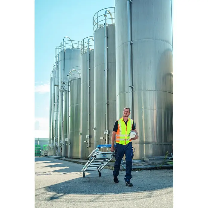Zarges Teleskopleiter fahrbar gebördelte Teleskop Plattformleiter ZAP SAFEMASTER MAX S 3 Stufen - Anzahl Tritte: 3 Stk., Arbeitshöhe: 2.700 m, Belastbarkeit: 150 kg, Aluminium - Mann in Warnweste transportiert eine zusammengeklappte, silber-blaue Mehrzweckleiter auf Rädern in einem Industriegebiet.