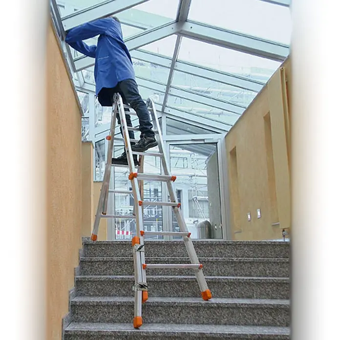 Layher Teleskopleiter Topic 1058 Teleskopleiter 4x3 Sprossen - Anzahl Tritte: 3 Stk., Arbeitshöhe: 3.850 m, Aluminium - Verstellbare Aluminiumleiter mit orangefarbenen Akzenten, flexibel auf einer Treppe zur Arbeit an einer Glasdecke aufgebaut.