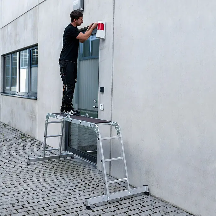 Munk Mehrzweckleiter Vielzweckleiter mit Nivello® Traverse 2x3 + 2x4 Sprossen - Anzahl Tritte: 4 Stk., Arbeitshöhe: 5.300 m, Aluminium - Mann auf einer silbernen, multifunktionalen Arbeitsplattform installiert Ausrüstung an einer Gebäudewand. Bietet sicheren erhöhten Zugang.