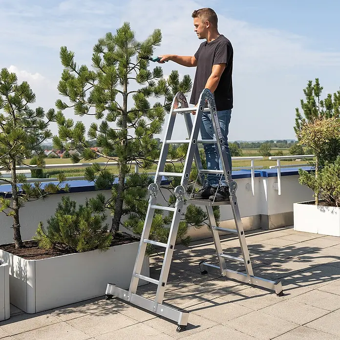 Munk Mehrzweckleiter Vielzweckleiter mit Nivello® Traverse 4x4 Sprossen - Anzahl Tritte: 4 Stk., Arbeitshöhe: 5.850 m, Aluminium - Silberne Aluminium-Mehrzweckleiter in Gebrauch. Ein Mann steht darauf und schneidet einen Baum auf einer sonnigen Terrasse.