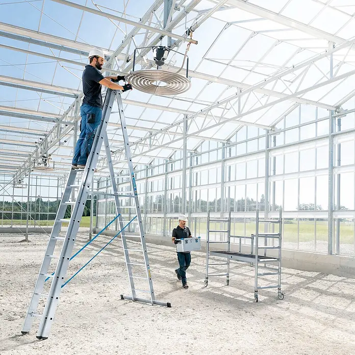 Zarges Mehrzweckleiter Skymaster X Mehrzweckleiter 3x9 Sprossen - Anzahl Tritte: 9 Stk., Arbeitshöhe: 7.450 m, Aluminium - Mehrzweck-Aluminiumleiter, von einem Arbeiter zur Installation von Geräten in einem Gewächshaus verwendet, mit Rollgerüst in der Nähe.