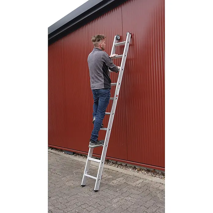 Euroline Mehrzweckleiter Premium 30777 Stufen Mehrzweckleiter mit Wandlaufrollen 3-teilig 3x8 Stufen - Anzahl Tritte: 8 Stk., Arbeitshöhe: 6.500 m, Belastbarkeit: 150 kg, Aluminium - Silberne Aluminiumleiter an roter Wand, ein Mann steigt hinauf.