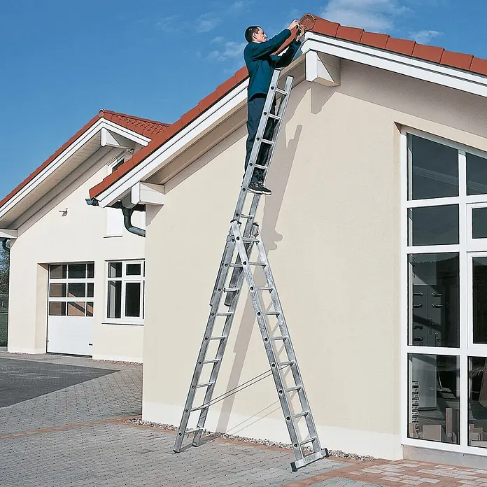 Zarges Mehrzweckleiter Skymaster DX Mehrzweckleiter 3x11 Sprossen - Anzahl Tritte: 11 Stk., Arbeitshöhe: 7.900 m, Aluminium - Silberne Aluminium-Mehrzweckleiter im Einsatz, ausgefahren um das Dach eines Hauses zu erreichen, wo ein Mann arbeitet.