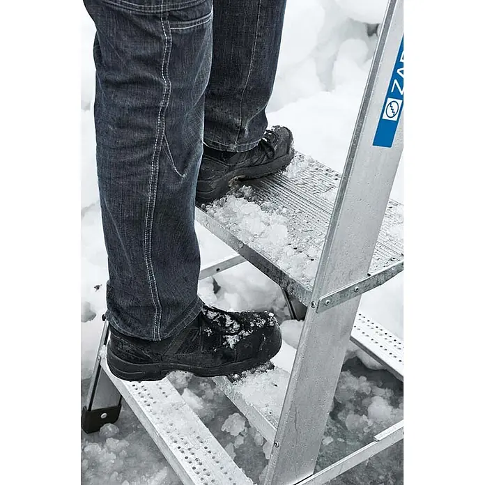 Zarges Arbeitsplattform Arbeitspodest Seventec RC SP 5 Stufen - Anzahl Tritte: 5 Stk., Arbeitshöhe: 3.080 m, Aluminium - Füße einer Person auf einer silbernen Leiter mit Schnee und Eis, zeigt sicheren Einsatz bei Winterbedingungen.
