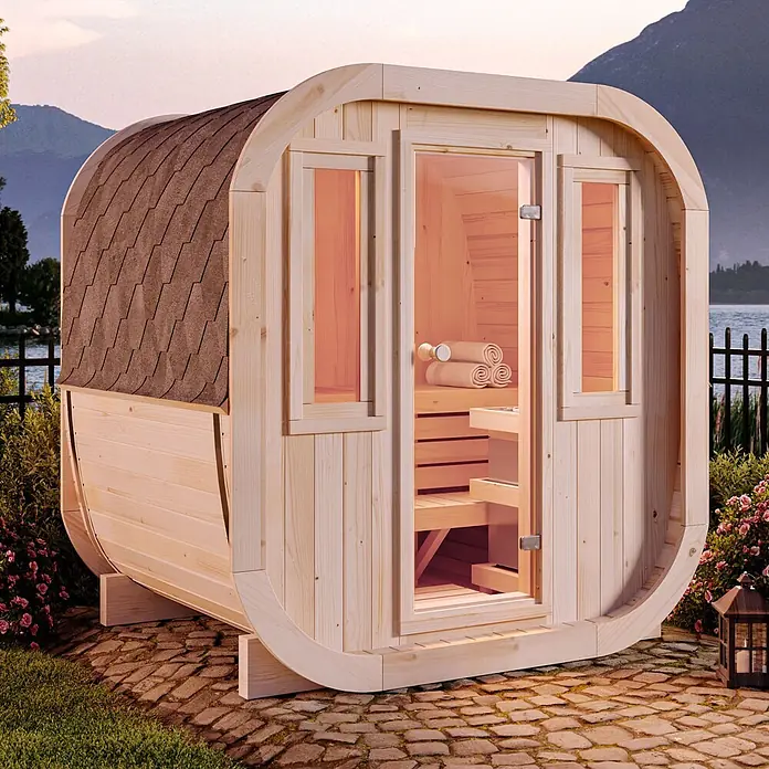 FinnTherm Fasssauna ScandiCube Mini XS - Holz, L x B x H: 166 x 21 x 214 cm - Fasssauna aus hellem Holz mit Schindeldach, auf einer Terrasse an einem See gelegen.