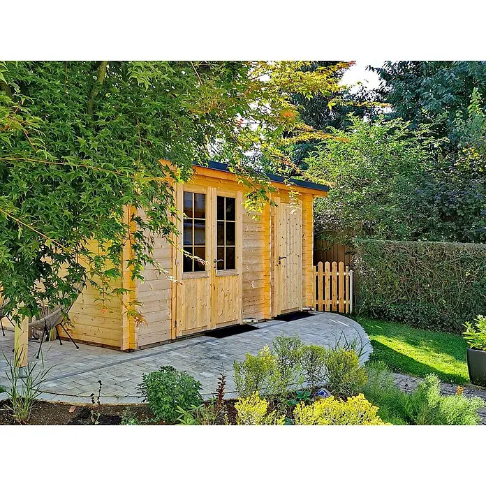 Fjordholz Gartenhaus Carin 44 C +SD - Außenmaß inkl. Dachüberstand (B x T): 606 x 320 cm, Holz, Kiefer/Pinie Imprägnierung - Helles Holzgartenhaus mit Fenstern und Tür auf einer gepflasterten Terrasse, umgeben von üppigem Grün.