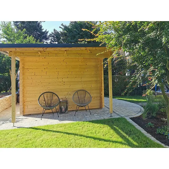 Fjordholz Gartenhaus Carin 44 C +SD - Außenmaß inkl. Dachüberstand (B x T): 606 x 320 cm, Holz, Kiefer/Pinie Imprägnierung - Heller Holzschuppen mit schwarzem Dach, der einen geschützten Bereich mit zwei Stühlen und einer gepflasterten Terrasse bietet.