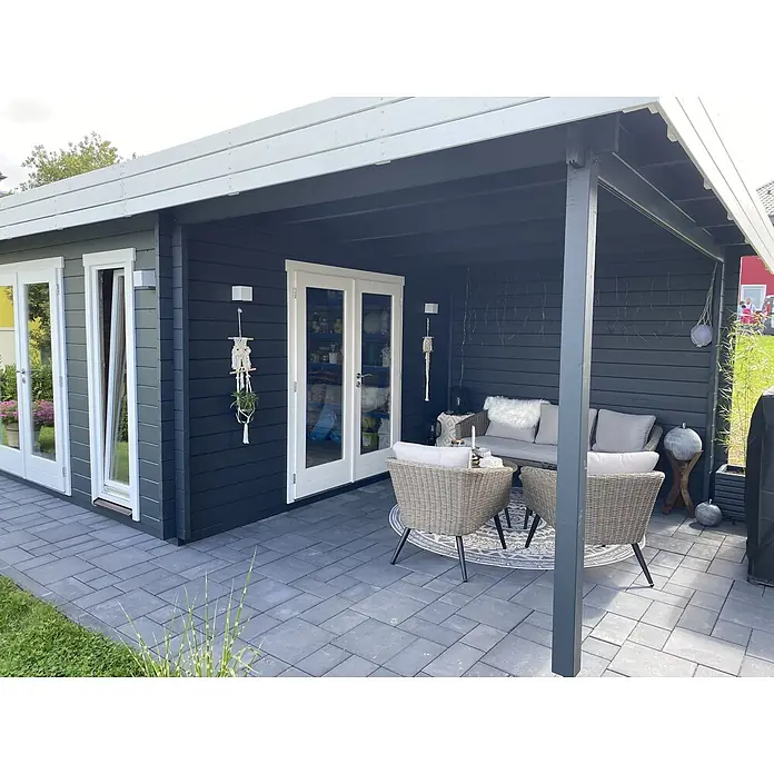 Fjordholz Gartenhaus Ronya 44 Farbbehandlung Carbongrau - Außenmaß inkl. Dachüberstand (B x T): 632 x 360 cm, Holz, Naturbelassen - Moderne Außenterrasse mit dunkelgrauem Gartenhaus und Korbmöbeln auf grauen Pflastersteinen, gemütlicher Entspannungsbereich.