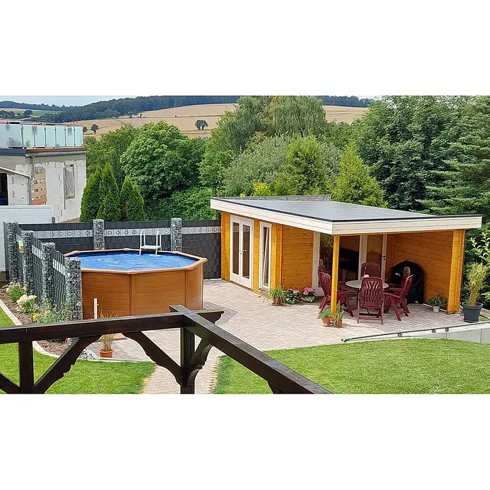 Fjordholz Gartenhaus Ronya 44 Farbbehandlung Carbongrau - Außenmaß inkl. Dachüberstand (B x T): 632 x 360 cm, Holz, Naturbelassen - Holzfarbenes Gartenhaus mit überdachter Terrasse und roten Möbeln, neben einem Aufstellpool in einem gepflegten Garten.