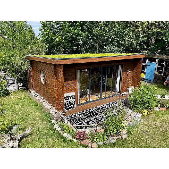 Fjordholz Gartenhaus - Außenmaß inkl. Dachüberstand (B x T): 597 x 465 cm, Holz, Grau Imprägnierung - Modernes Gartenhaus aus Holz mit Gründach, großen Glastüren und dekorativem Gitter auf der Holzterrasse in einem üppigen Garten.