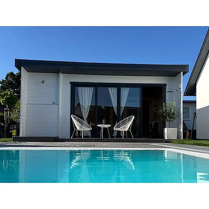 Fjordholz Gartenhaus - Außenmaß inkl. Dachüberstand (B x T): 597 x 465 cm, Holz, Grau Imprägnierung - Modernes weißes Poolhaus mit dunklen Akzenten, Glasschiebetüren und kleiner Terrasse mit weißen Stühlen neben einem blauen Pool.
