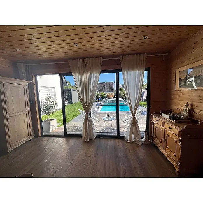 Fjordholz Gartenhaus - Außenmaß inkl. Dachüberstand (B x T): 597 x 465 cm, Holz, Grau Imprägnierung - Holzzimmer mit großen Glastüren, Blick auf sonnigen Garten mit Pool und Terrasse.