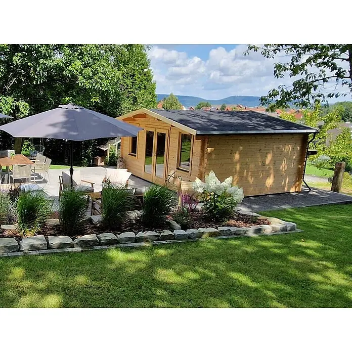 Fjordholz Gartenhaus Agnes 44 Farbbehandlung Lichtgrau - Außenmaß inkl. Dachüberstand (B x T): 500 x 400 cm, Holz, Naturbelassen - Holzgartenhaus mit Terrasse, Sitzgelegenheiten und Sonnenschirm, umgeben von Gartenpflanzen.