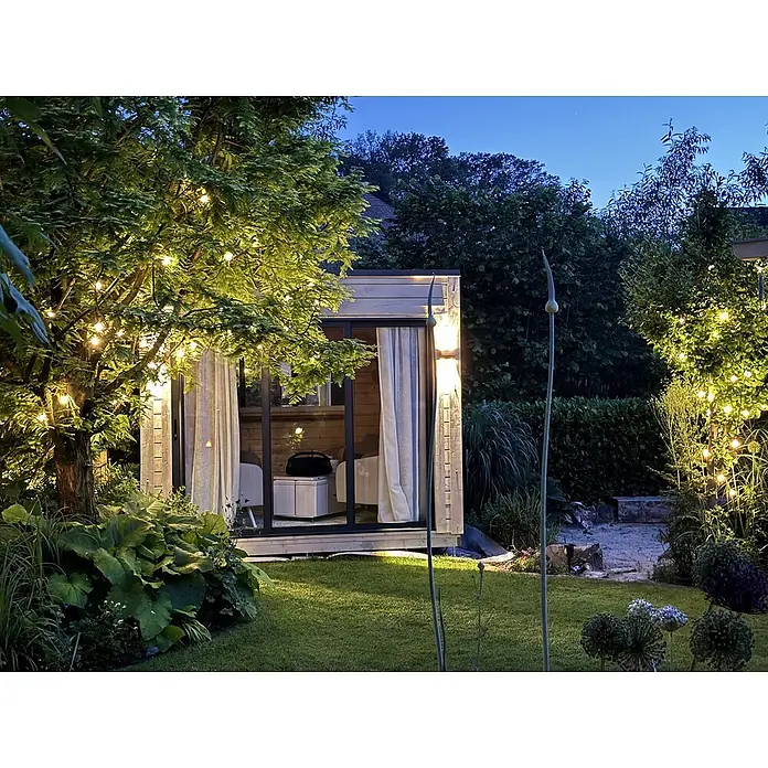 Fjordholz Gartenhaus Q-BIC Alu 70 G 6mm Einfachglas Grau Imprägnierung - Außenmaß inkl. Dachüberstand (B x T): 300 x 300 cm, Holz, Naturbelassen - Moderne Gartenhütte aus hellem Holz mit Glastüren, nachts in einem gemütlichen Garten beleuchtet.