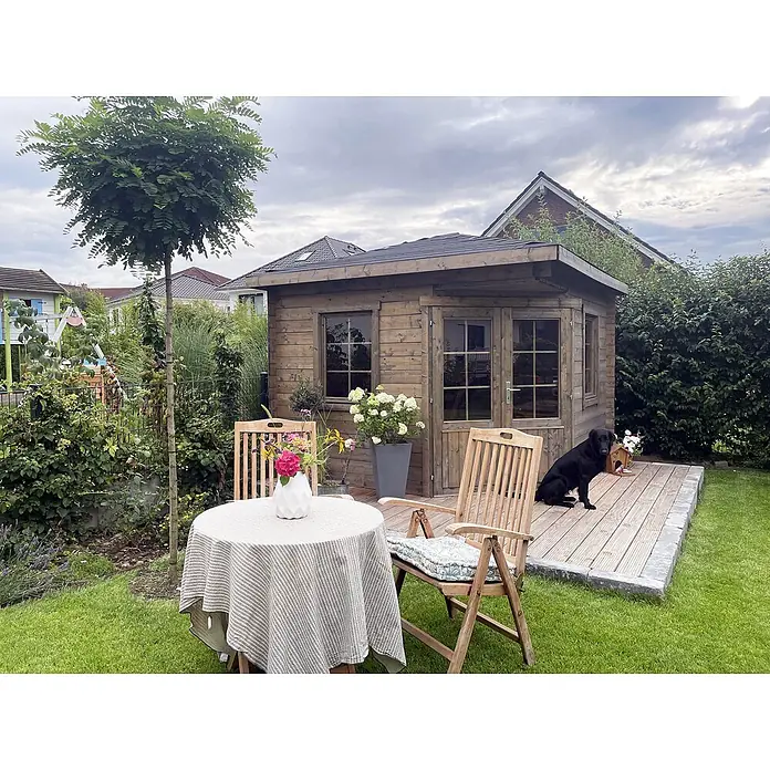 Fjordholz Gartenhaus Evelyn 3A - Außenmaß inkl. Dachüberstand (B x T): 320 x 320 cm, Holz, Naturbelassen - Rustikaler Holz-Gartenschuppen mit Fenstern, kleiner Terrasse und angrenzender Outdoor-Essgruppe, mit einem schwarzen Hund.