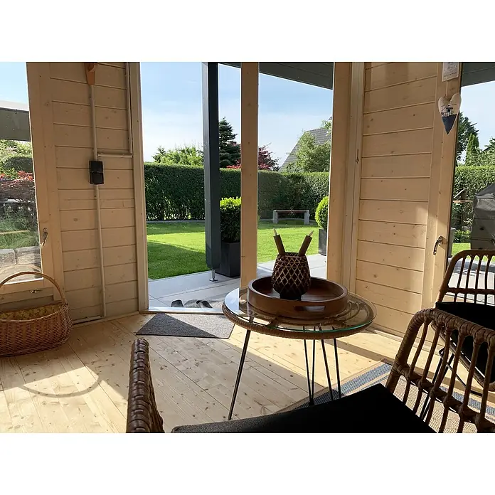 Fjordholz Gartenhaus Nora 44 H Modern - Außenmaß inkl. Dachüberstand (B x T): 740 x 320 cm, Holz, Farbbehandlung Lichtgrau - Ein Beistelltisch mit Glasplatte, Holztablett und dunkler Vase, umgeben von Rattanstühlen in einem sonnigen Raum mit Gartenblick.