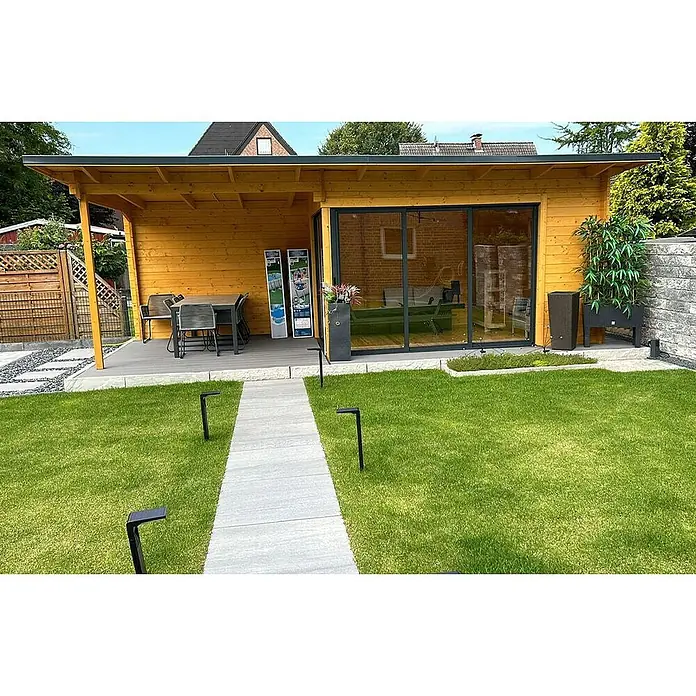 Fjordholz Gartenhaus Starla 44 D - Außenmaß inkl. Dachüberstand (B x T): 731,6 x 325 cm, Holz, Naturbelassen - Modernes Gartenhaus aus Holz mit überdachter Terrasse, Glasschiebetüren und Gartenmöbeln in einem grünen Garten mit Pflasterweg.