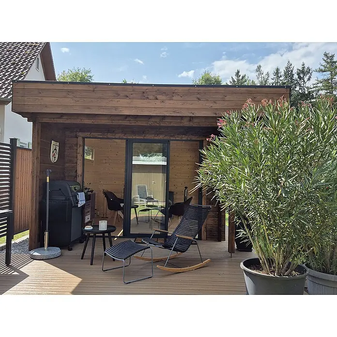 Fjordholz Gartenhaus - Außenmaß inkl. Dachüberstand (B x T): 448 x 398 cm, Holz, Grau Imprägnierung - Schwarzer Outdoor-Schaukelstuhl und passender Fußhocker auf Holzterrasse, als entspannter Sitzbereich am Gartenhaus.