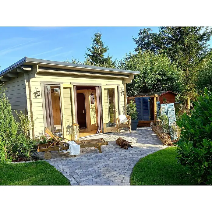 Fjordholz Gartenhaus Jacob 44 E - Außenmaß inkl. Dachüberstand (B x T): 471,2 x 351,2 cm, Holz, Grau Imprägnierung - Hellgelbes Gartenhaus aus Holz mit Glastüren, das sich zu einer gepflasterten Terrasse mit Gartenmöbeln und einem Hund öffnet.