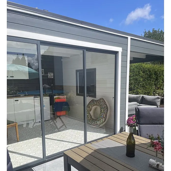 Fjordholz Gartenhaus Q-BIC Alu 44 A + SD 6mm Einfachglas - Außenmaß inkl. Dachüberstand (B x T): 680 x 380 cm, Holz, ohne Imprägnierung - Moderner Außenwohnbereich mit grauen Glasschiebetüren, die sich zu einer Terrasse mit kleiner Küche und Sitzgelegenheiten öffnen.