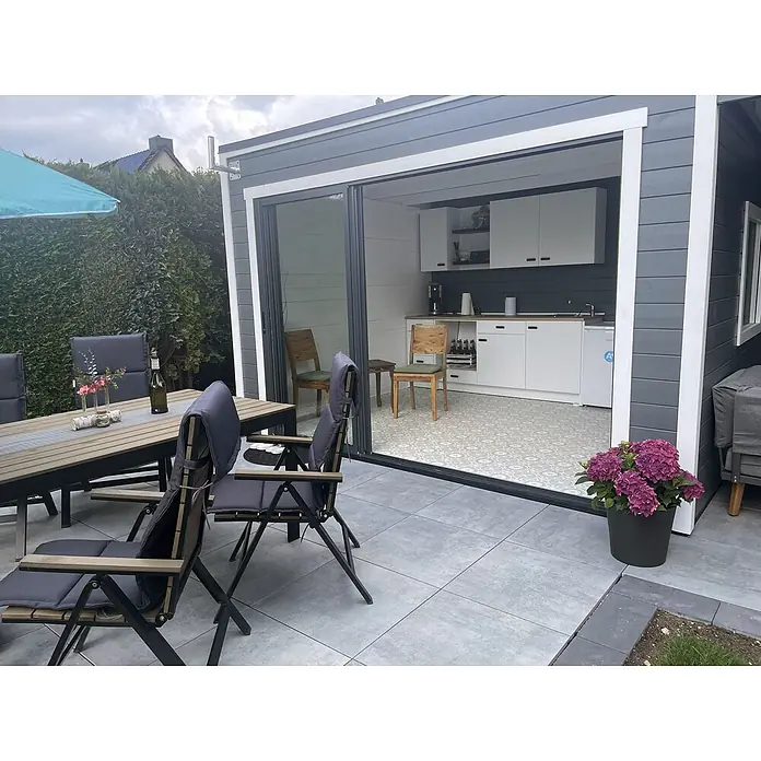 Fjordholz Gartenhaus Q-BIC Alu 44 A + SD 6mm Einfachglas - Außenmaß inkl. Dachüberstand (B x T): 680 x 380 cm, Holz, ohne Imprägnierung - Eine Outdoor-Essgruppe mit Holztisch und dunkelgrauen gepolsterten Stühlen auf einer grauen Fliesenterrasse neben einem Gartenhaus.