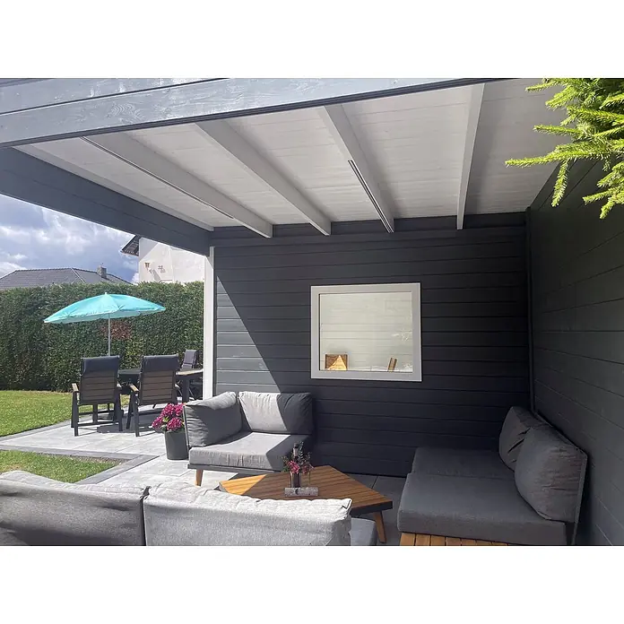 Fjordholz Gartenhaus Q-BIC Alu 44 A + SD 6mm Einfachglas - Außenmaß inkl. Dachüberstand (B x T): 680 x 380 cm, Holz, ohne Imprägnierung - Moderne dunkelgraue Pergola mit weißer Decke, die einen schattigen Outdoor-Loungebereich mit grauen Kissen und Holztischen bietet.