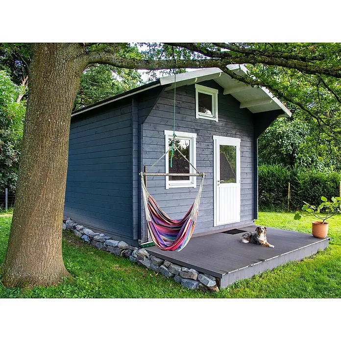Fjordholz Gartenhaus Sönke 44 B - Außenmaß inkl. Dachüberstand (B x T): 388 x 488 cm, Holz, ohne Imprägnierung - Ein dunkelgraues Holzgartenhaus mit weißen Akzenten, mit einer bunten Hängematte am Baum und einem Hund auf der Holzterrasse.