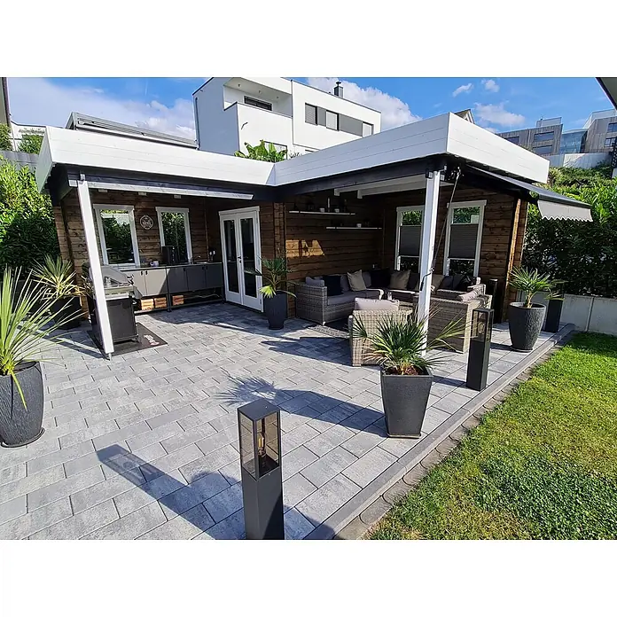 Fjordholz Gartenhaus Fred 44 Grau Imprägnierung - Außenmaß inkl. Dachüberstand (B x T): 573,2 x 573,2 cm, Holz, Naturbelassen - Outdoor-Wohnbereich mit Holzgartenhaus, überdachter Terrasse, Außenküche und Lounge auf grauen Rechteckpflastersteinen.