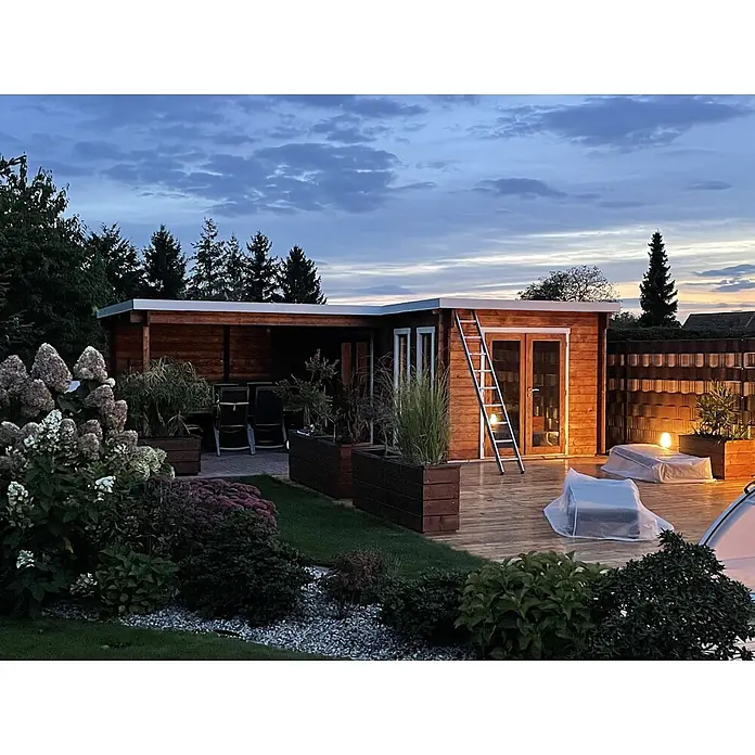 Fjordholz Gartenhaus Luisa 44 - Außenmaß inkl. Dachüberstand (B x T): 680 x 480 cm, Holz, Naturbelassen - Ein modernes Gartenhaus aus Holz mit Flachdach, großzügiger Holzterrasse, abgedeckten Möbeln und Pflanzkübeln, beleuchtet in der Dämmerung.