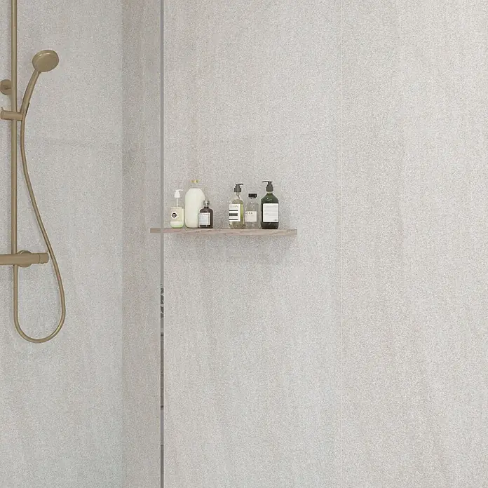 Wetwall Wandpaneel Elite - 16 x 242 cm, Moonlit Sand, Abgerundete Kante - Hellbeige gesprenkelte Wandfliesen in einer modernen Dusche, mit einem goldenen Duschkopf und einem Holzregal mit Toilettenartikeln.