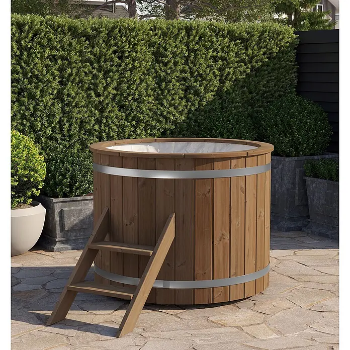 Tuindeco Eisbad - Ø x H: 133 x 109 cm, Natur - Holzbadefass mit silbernen Bändern und Leiter auf einer Terrasse, umgeben von Hecken und Pflanzgefäßen.