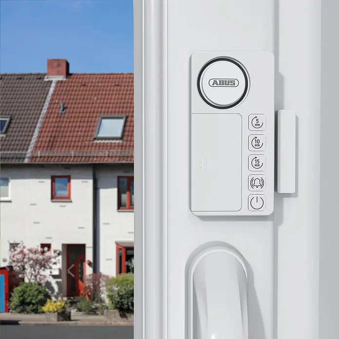 Abus Alarmanlage Time2Close Window Countdown Alarm - Weiß, 15 x 43 x 85 mm - Weißes ABUS Sicherheitssystem am Türrahmen mit Zeit- und Alarmeinstellungen, Häuser im Hintergrund.