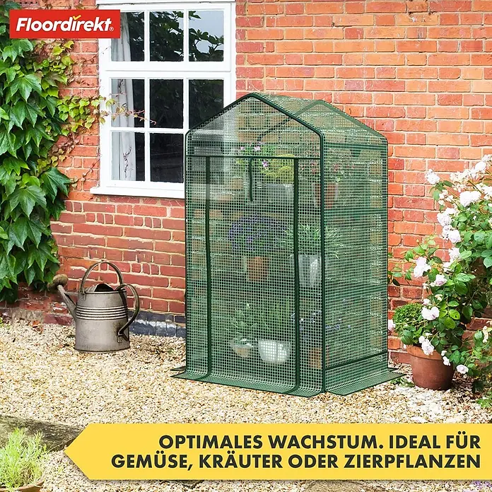 Floordirekt Folien-Gewächshaus Bloom - 49 x 69 x 93 cm, 2 Etagen, aufrollbare Tür mit Reißverschluss - Grünes Netz-Mini-Gewächshaus mit Pflanzen auf Kies im Garten vor einem Backsteinhaus.