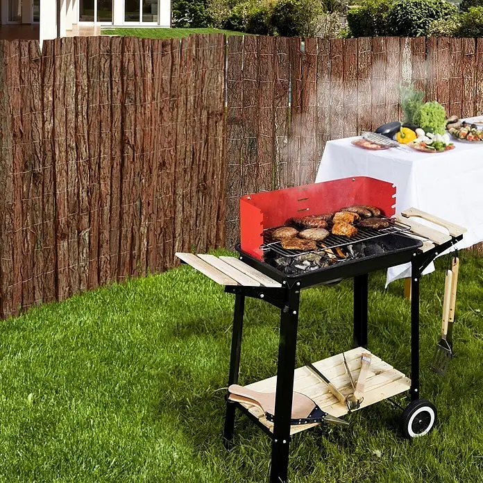 Balkonsichtschutz Rindenmatte - 300 x 150 cm, Braun - Schwarzer und roter Grill mit Fleisch, Holzablagen und Werkzeugen, neben Sichtschutzzaun und Tisch.